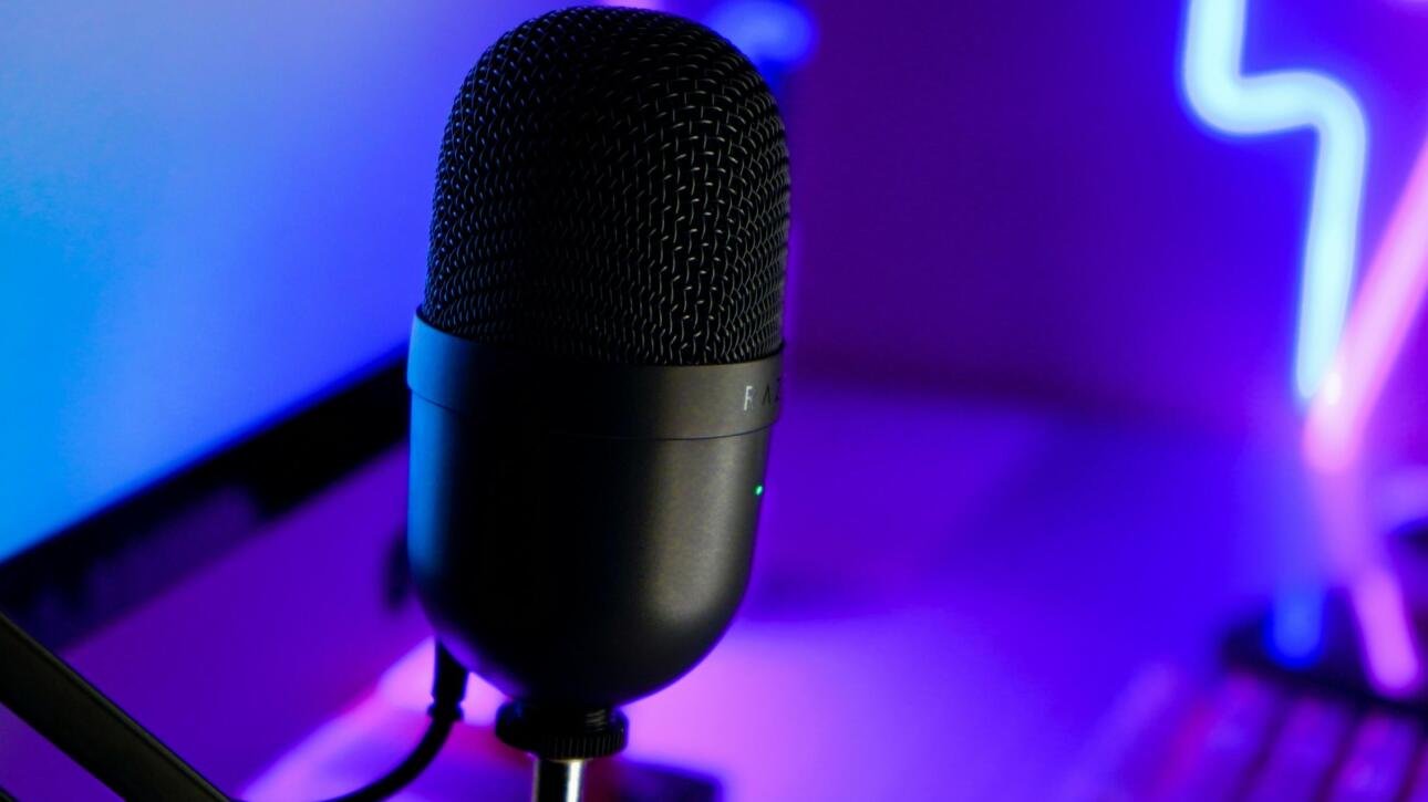 A professional black condenser microphone with a metal mesh grille, positioned in a vibrant neon-lit streaming setup with purple and blue lighting. Ideal for live streaming, podcasting, and gaming.