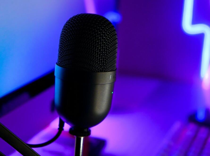 A professional black condenser microphone with a metal mesh grille, positioned in a vibrant neon-lit streaming setup with purple and blue lighting. Ideal for live streaming, podcasting, and gaming.