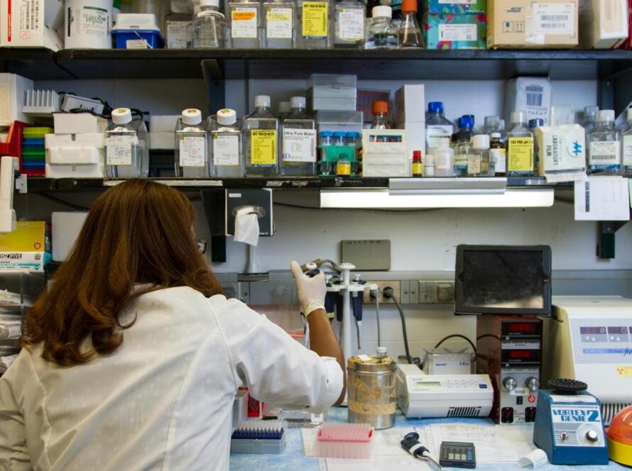 A laboratory technician in a white coat conducting STD testing in a research lab with various medical supplies and equipment on shelves.
